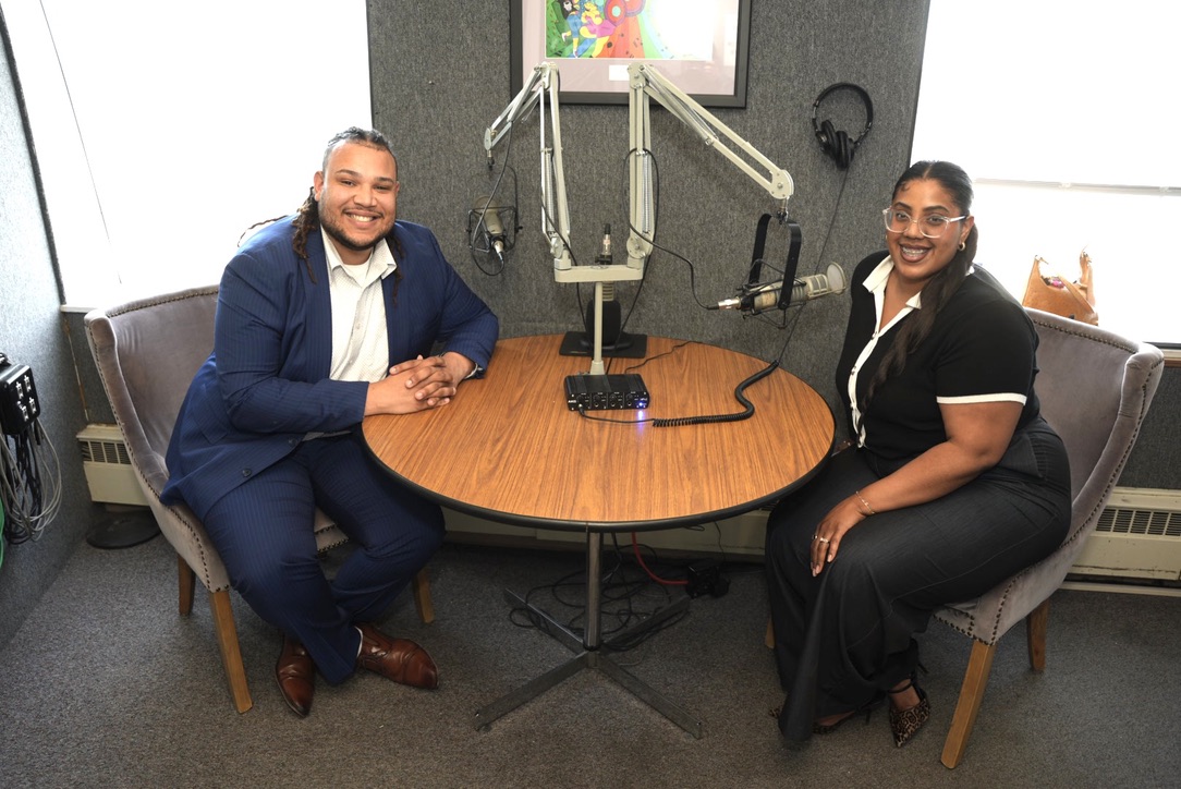 Two people smiling across a radio studio table with microphones