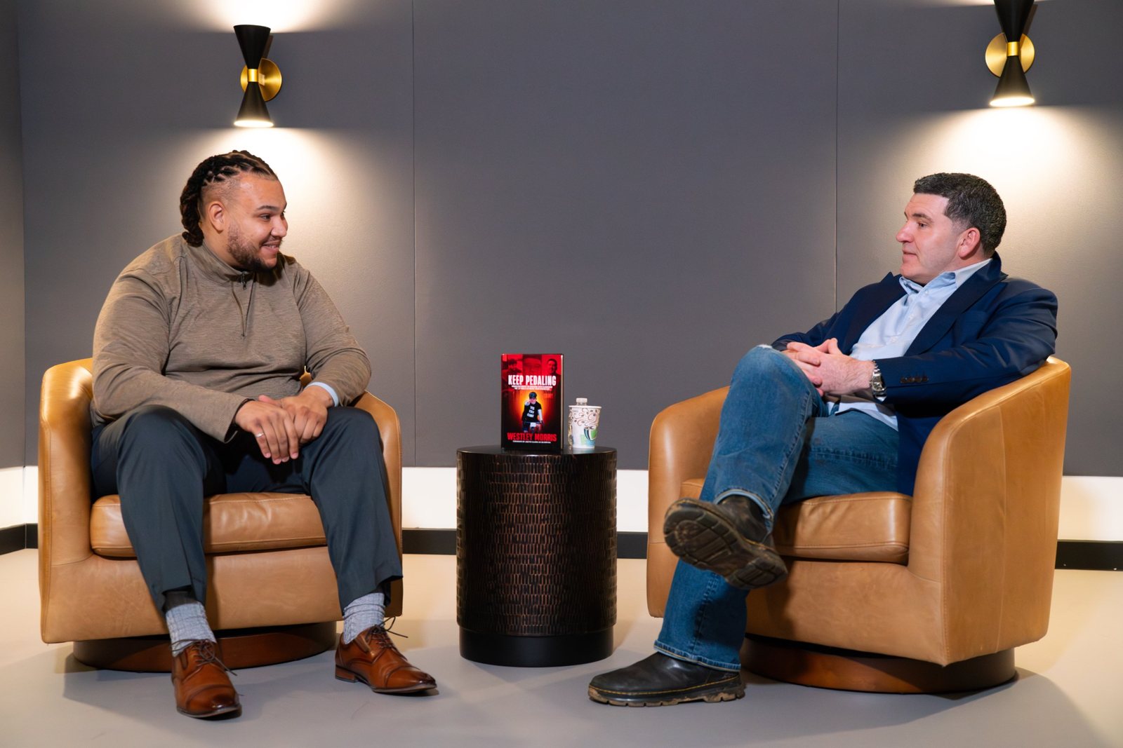 Carter McKinney interviewing a guest in the studio, seated in leather chairs with a book on the table between them